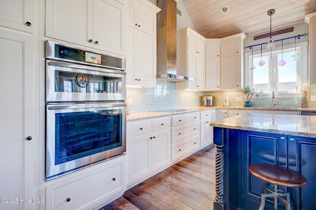 kitchen with tasteful backsplash, light stone counters, stainless steel double oven, wall chimney range hood, and white cabinets