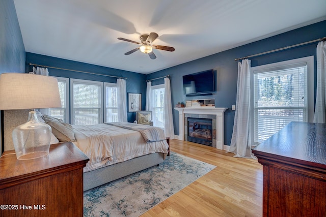 bedroom featuring ceiling fan, a tiled fireplace, light hardwood / wood-style floors, and multiple windows