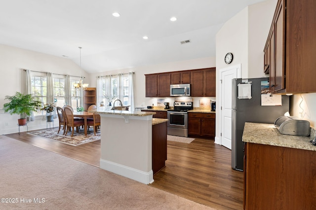 kitchen with lofted ceiling, appliances with stainless steel finishes, a kitchen island with sink, light stone countertops, and decorative light fixtures