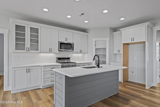 kitchen with sink, appliances with stainless steel finishes, a kitchen island with sink, white cabinets, and light wood-type flooring