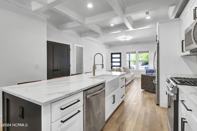 kitchen with an island with sink, beamed ceiling, stainless steel appliances, light stone countertops, and white cabinets