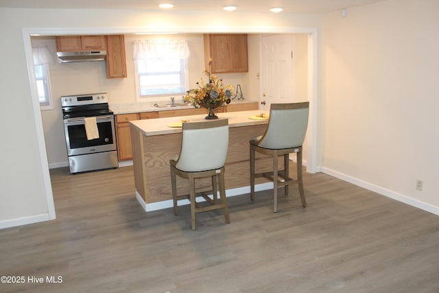 kitchen featuring light countertops, stainless steel range with electric stovetop, under cabinet range hood, a breakfast bar area, and wood finished floors