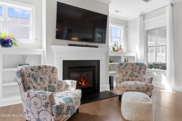 living area featuring wood finished floors, a glass covered fireplace, visible vents, and crown molding