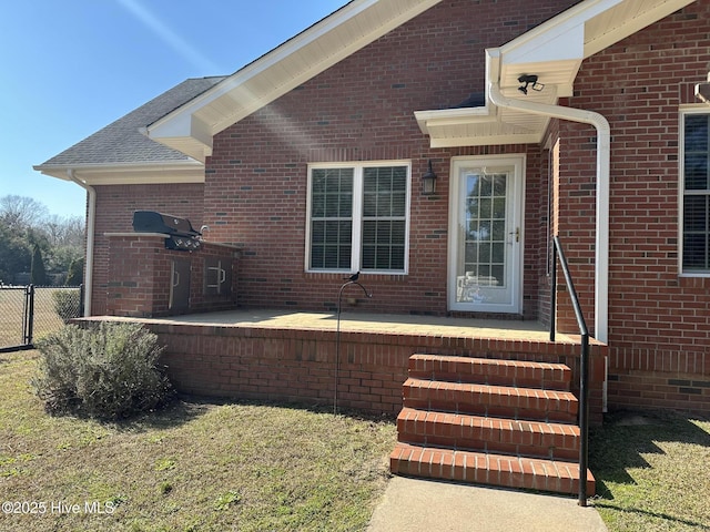 property entrance featuring brick siding, a patio area, roof with shingles, and fence