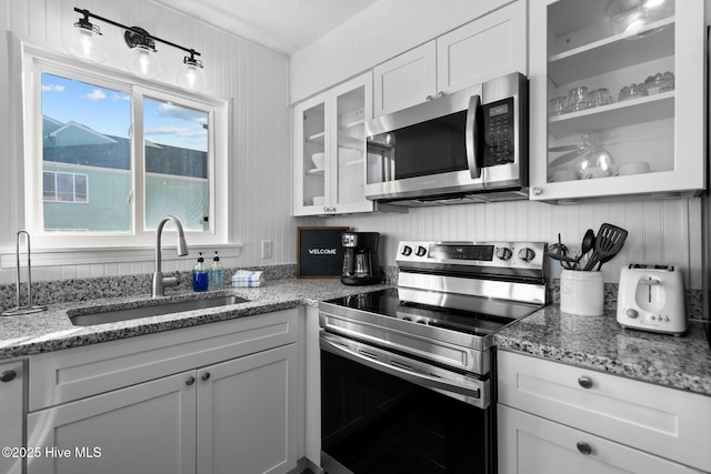 kitchen with light stone counters, a sink, white cabinetry, appliances with stainless steel finishes, and glass insert cabinets