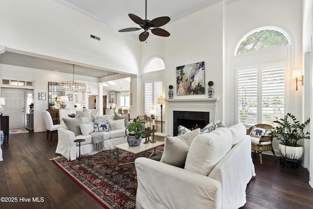 living area featuring ceiling fan, a warm lit fireplace, dark wood-style flooring, a towering ceiling, and crown molding
