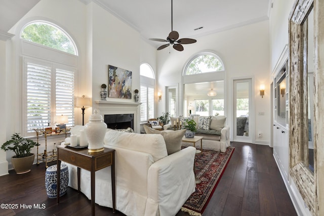 living room with a fireplace, a ceiling fan, dark wood-style flooring, a high ceiling, and ornamental molding