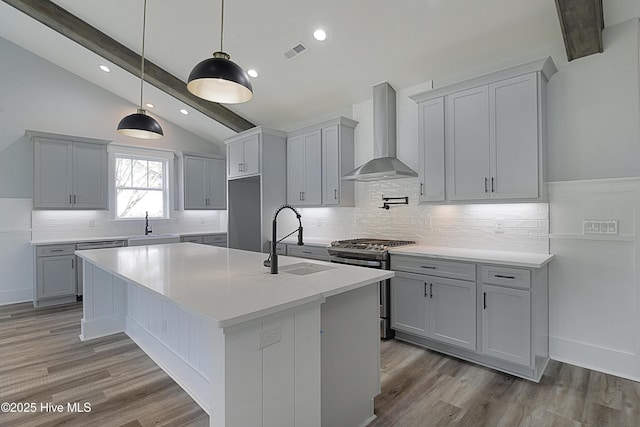 kitchen featuring sink, vaulted ceiling with beams, wall chimney range hood, gas stove, and a center island with sink