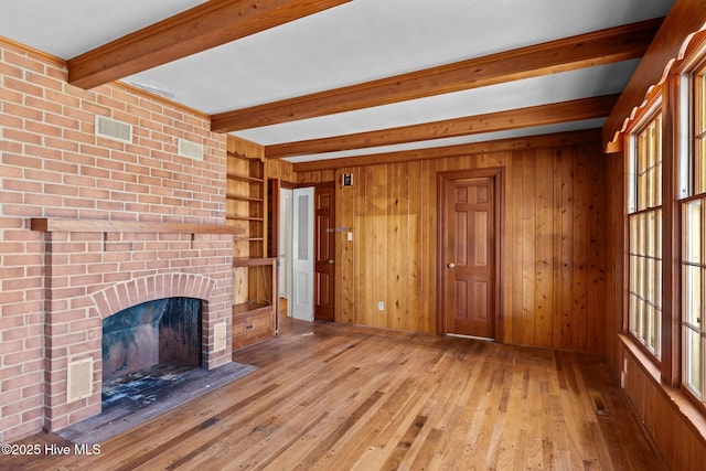 unfurnished living room with a brick fireplace, built in shelves, beamed ceiling, wood walls, and light hardwood / wood-style floors