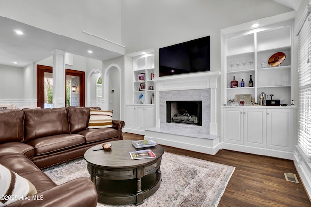 living room with dark wood-type flooring, built in shelves, ornate columns, a towering ceiling, and a premium fireplace