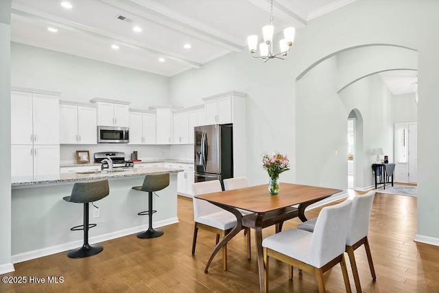 dining room with sink, light hardwood / wood-style flooring, a towering ceiling, beamed ceiling, and a chandelier