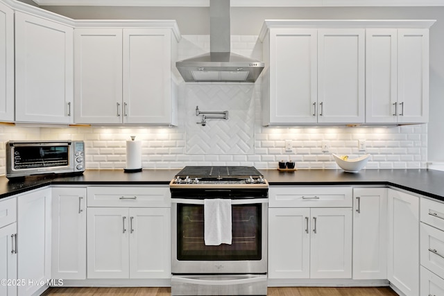 kitchen featuring stainless steel gas range oven, white cabinetry, dark countertops, wall chimney exhaust hood, and tasteful backsplash