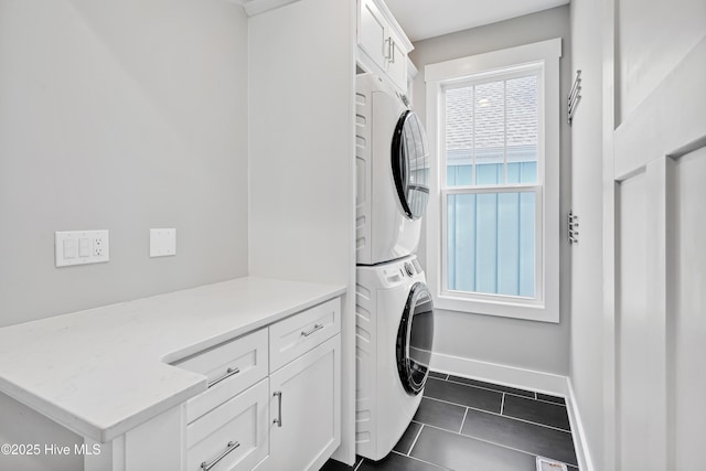 washroom featuring baseboards, cabinet space, stacked washer and clothes dryer, and dark tile patterned flooring