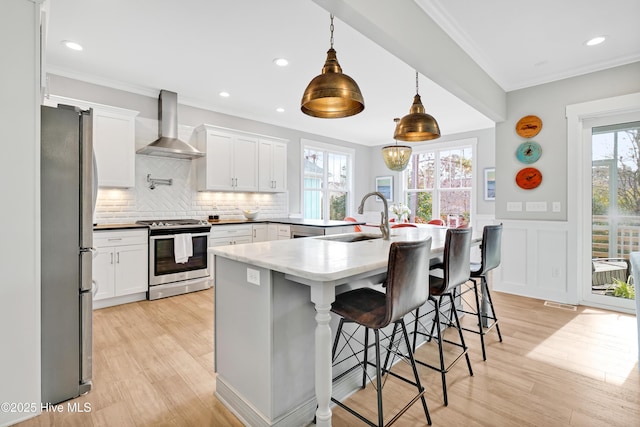 kitchen featuring ornamental molding, a sink, appliances with stainless steel finishes, wall chimney exhaust hood, and light wood-type flooring