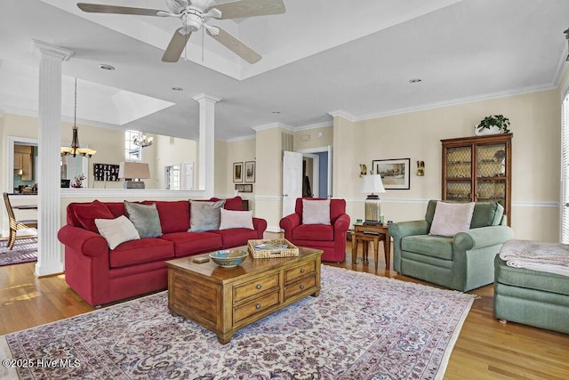 living room with ornate columns, ceiling fan with notable chandelier, a raised ceiling, crown molding, and light wood-type flooring