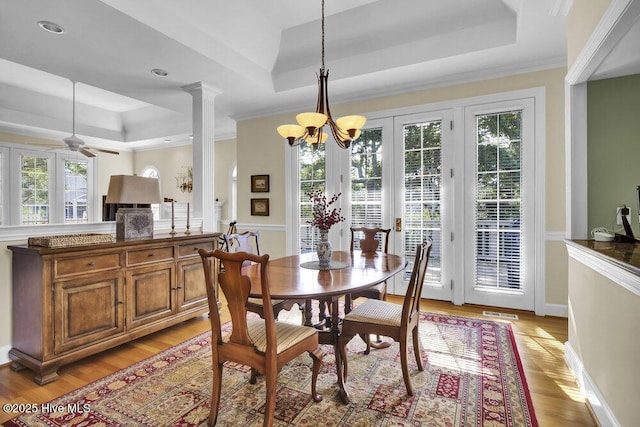 dining space featuring ornate columns, ceiling fan with notable chandelier, light wood-type flooring, and a tray ceiling