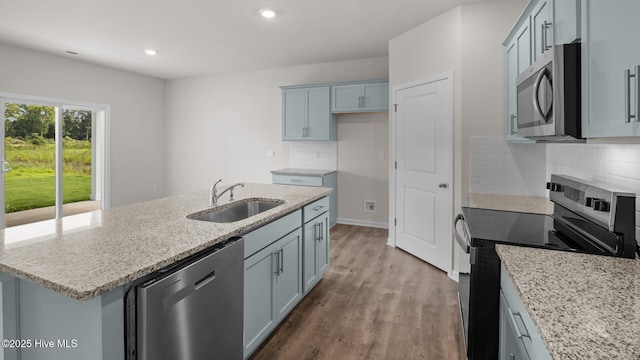 kitchen with appliances with stainless steel finishes, hardwood / wood-style floors, sink, a kitchen island with sink, and light stone countertops