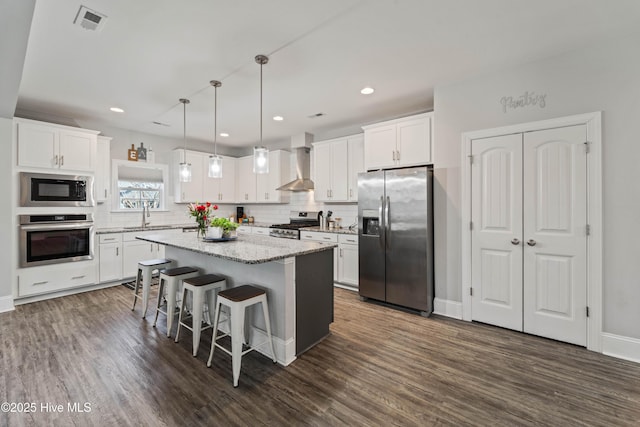 kitchen featuring appliances with stainless steel finishes, decorative light fixtures, white cabinetry, a center island, and wall chimney range hood