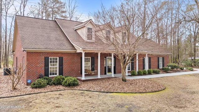 new england style home with brick siding, covered porch, central AC, and a shingled roof