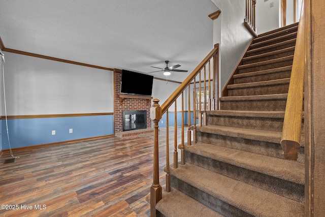 staircase featuring a textured ceiling, wood finished floors, baseboards, ornamental molding, and a brick fireplace