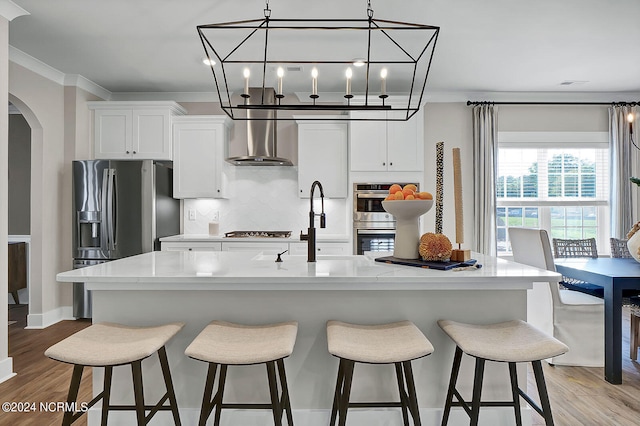 kitchen featuring stainless steel appliances, a kitchen island with sink, white cabinets, and wall chimney exhaust hood