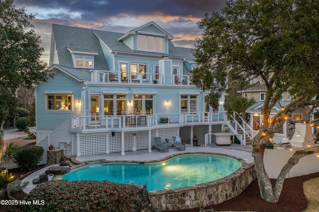 back of property at dusk featuring a shingled roof, a patio area, stairs, an outdoor pool, and a balcony