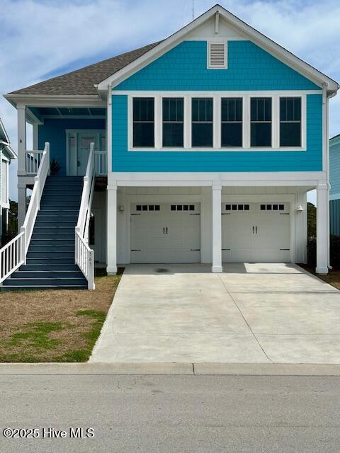 coastal home featuring driveway, stairway, roof with shingles, an attached garage, and a porch