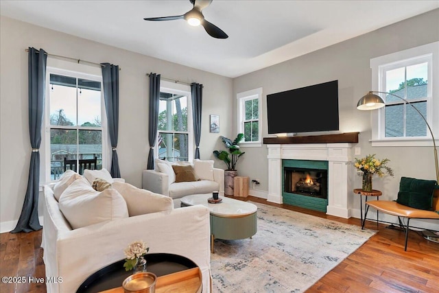 living room featuring a lit fireplace, ceiling fan, baseboards, and wood finished floors