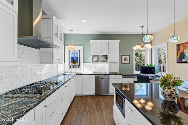 kitchen featuring wall chimney exhaust hood, stainless steel appliances, dark stone counters, white cabinetry, and hanging light fixtures