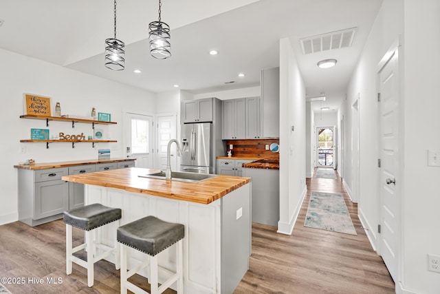 kitchen featuring sink, stainless steel fridge, wooden counters, gray cabinetry, and an island with sink