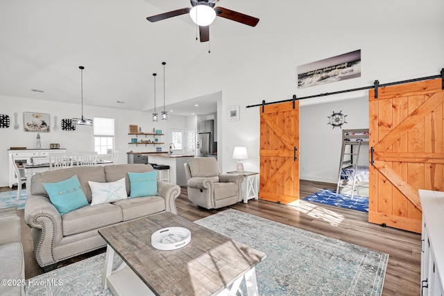 living room with ceiling fan, wood-type flooring, a barn door, and high vaulted ceiling