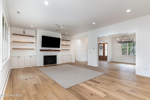 unfurnished living room with built in shelves, a large fireplace, ceiling fan with notable chandelier, and light hardwood / wood-style flooring