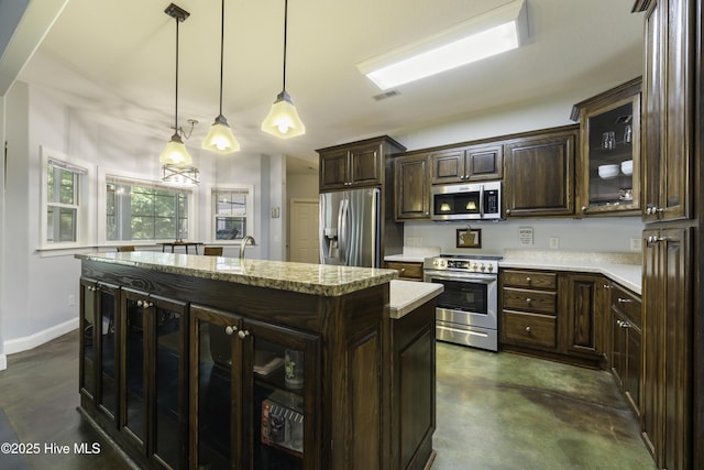 kitchen with a kitchen island with sink, dark brown cabinets, stainless steel appliances, light stone countertops, and decorative light fixtures