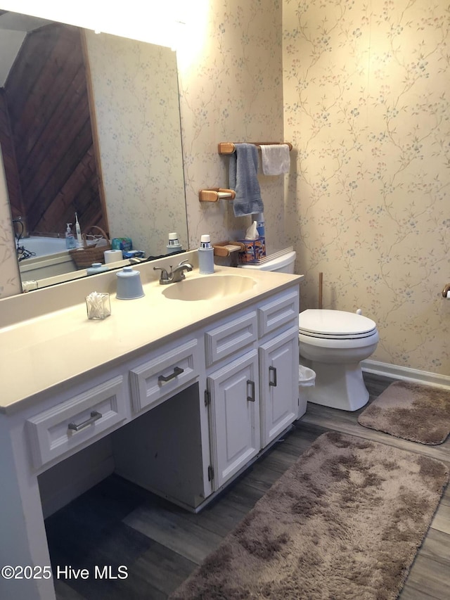 bathroom with toilet, wood finished floors, wallpapered walls, baseboards, and vanity