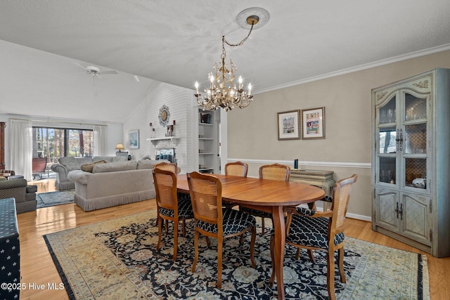 dining space with crown molding, a fireplace, light wood finished floors, lofted ceiling, and ceiling fan with notable chandelier