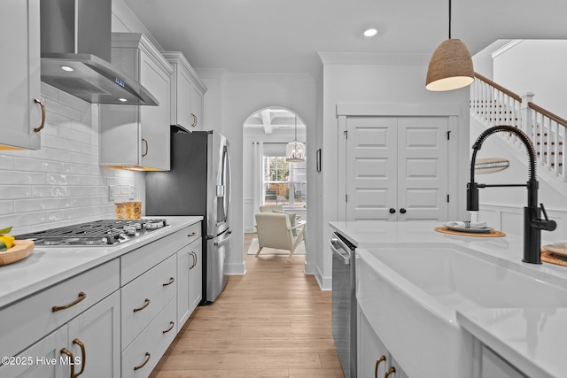 kitchen with appliances with stainless steel finishes, sink, hanging light fixtures, wall chimney range hood, and light hardwood / wood-style flooring