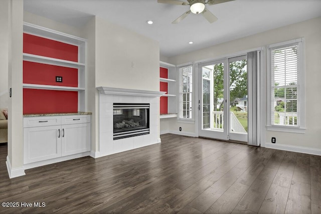 unfurnished living room with a ceiling fan, dark wood-style flooring, built in features, a tile fireplace, and baseboards