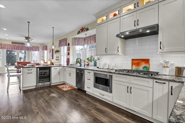 kitchen with hanging light fixtures, stainless steel appliances, beverage cooler, kitchen peninsula, and dark stone counters