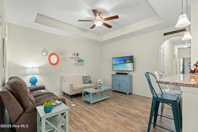 living area with arched walkways, ceiling fan, baseboards, light wood finished floors, and a tray ceiling