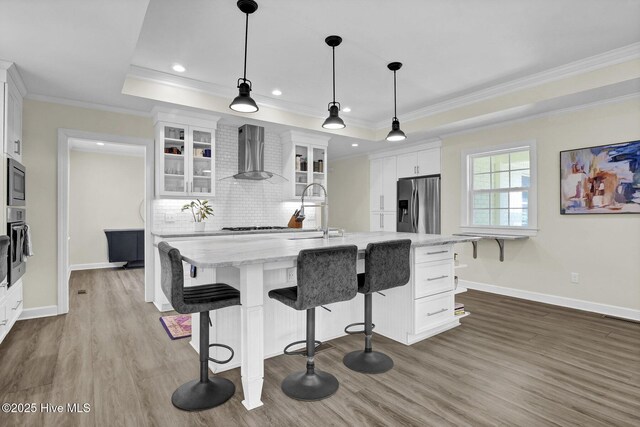 kitchen featuring wall chimney range hood, a tray ceiling, stainless steel appliances, white cabinetry, and a sink