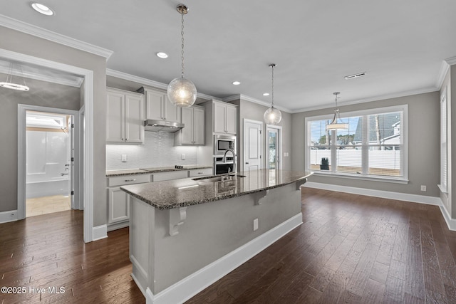 kitchen featuring an island with sink, sink, stainless steel microwave, dark stone countertops, and a kitchen bar