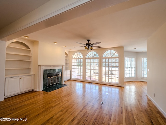 unfurnished living room with built in shelves, a fireplace, wood finished floors, a ceiling fan, and baseboards