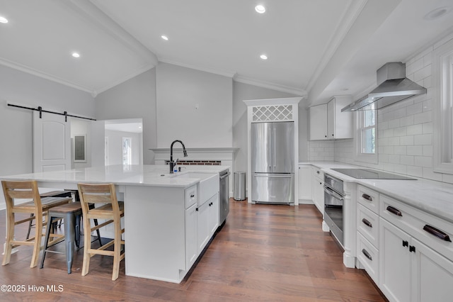 kitchen with a barn door, stainless steel appliances, white cabinets, wall chimney range hood, and an island with sink