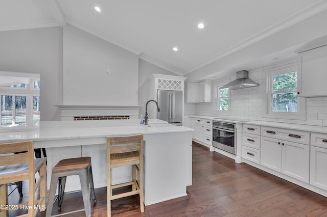 kitchen featuring a breakfast bar, white cabinetry, vaulted ceiling, appliances with stainless steel finishes, and wall chimney exhaust hood