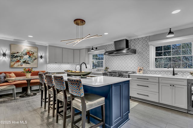 kitchen featuring a kitchen island with sink, a sink, light countertops, ornamental molding, and wall chimney range hood