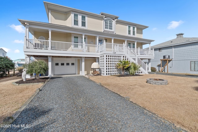 coastal inspired home with an outdoor fire pit, covered porch, a garage, stairs, and gravel driveway