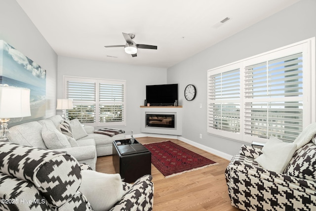 living room featuring visible vents, baseboards, a ceiling fan, a glass covered fireplace, and wood finished floors