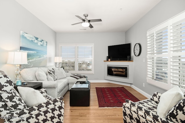 living area with a ceiling fan, a glass covered fireplace, baseboards, and light wood finished floors