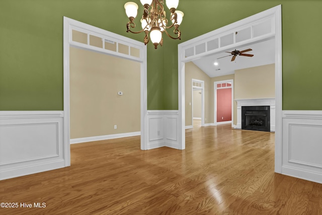 unfurnished living room featuring a wainscoted wall, a fireplace with flush hearth, vaulted ceiling, wood finished floors, and ceiling fan with notable chandelier