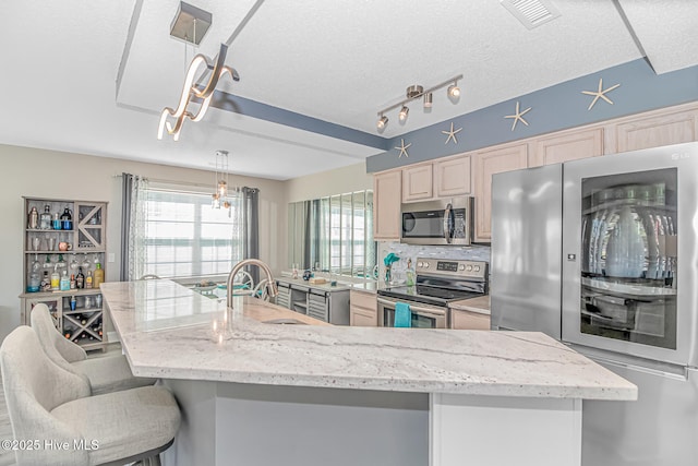 kitchen featuring light brown cabinetry, a textured ceiling, appliances with stainless steel finishes, an island with sink, and pendant lighting